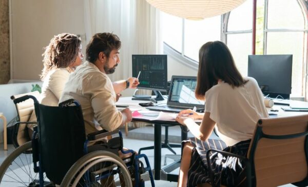mages of two females and a male sat at a desk looking at a laptop. The male is sat in a wheelchair